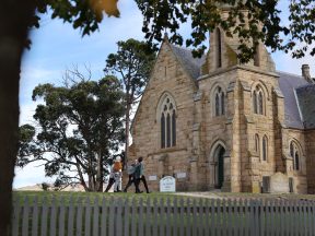 Photo of the Ross Uniting Church from a distance as a group of four friends approach its front door on a windy day.